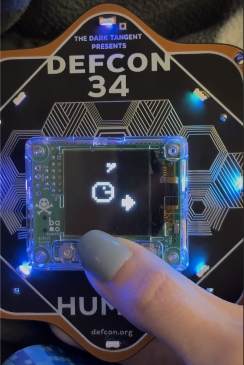 The DEFCON 34 badge with glowing LEDs around its edge, its OLED screen showing a pixel-art Tamagotchi while a finger presses one of the buttons below.